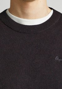Detailní záběr osoby v tmavém pleteném svetru přes bílém tričku s kulatým výstřihem, zobrazující výstřih a část oblasti hrudníku.