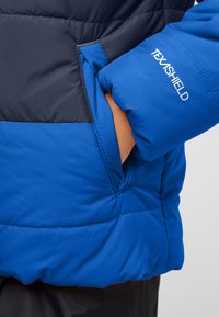 Giacca isolante blu con sezione superiore blu navy, design trapuntato e tasca laterale. Presenta il marchio "TEXASHIELD" sulla manica.