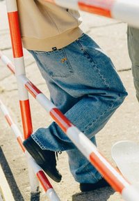 Luźne niebieskie dżinsy z żółtym logo na tylnym kieszeniu, w połączeniu z czarnym obuwiem, opierające się o czerwoną i białą barierę w paski.