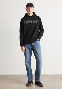 Felpa nera con logo, jeans blu e sneaker neri. La felpa ha una tasca frontale e un cappuccio con lacci, realizzata in cotone.