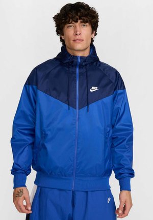 Veste Nike bleue avec capuche, fermeture éclair et section supérieure contrastante en bleu foncé. Fabriquée en matériau lisse et léger.