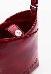 Piros bőrtáska sima, fényes felülettel. Ezüst színű patent zár és domború logó található az oldalán. Kompakt, struktúrált forma.