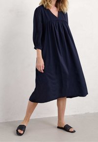 Vestido midi de corte holgado en azul marino con escote en V y mangas tres cuartos; cuenta con cintura fruncida y tejido suave. Sandalias deslizantes tejidas en negro.
