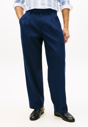 Άνδρας που φοράει μπλε-σκούρο υφασμάτινο παντελόνι, μαύρα δερμάτινα loafers και ανοικτό γαλάζιο πουκάμισο με ρίγες, με τα μανίκια σηκωμένα.