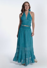 Un top a halter e una gonna lunga a strati in teal, con una vita arricciata e un tessuto leggero, che esalta il movimento e la fluidità.