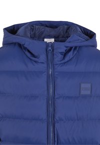 Mėlyna pūkinė striukė su gaubtu, su visa užtrauktuko uždanga, siuvinėta tekstūra ir įspaustu logotipu ant krūtinės.