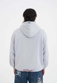 Felpa grigia chiara con cappuccio, maniche lunghe, texture morbida e dettaglio ricamato "Ed Hardy" sull'orlo, indossata con pantaloni in denim blu.