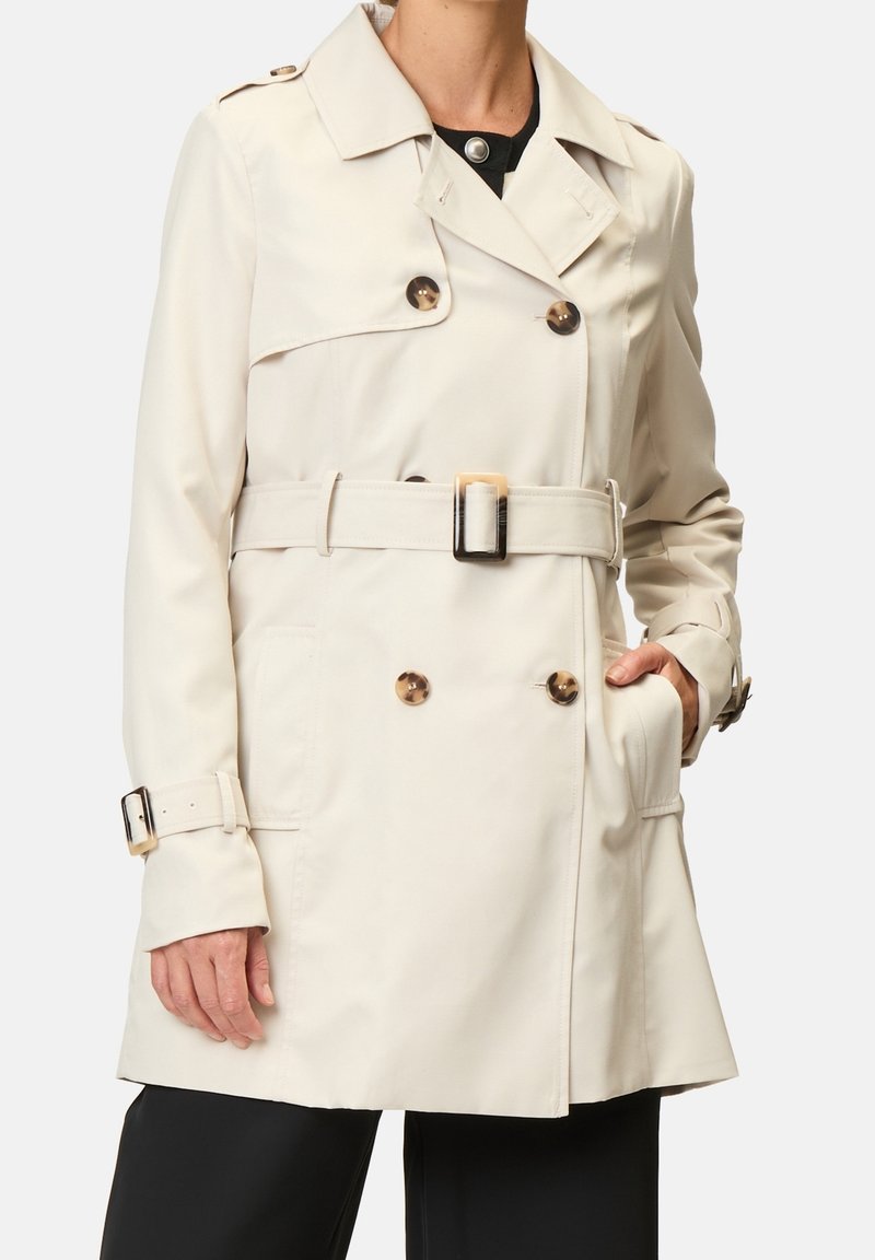 Trench-coat beige à boutons croisés, ceinturé à la taille, avec des poches latérales. Il comporte un col classique et des manches longues.