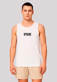 Weißes Baumwoll-Tanktop mit Rundhalsausschnitt, auf der Vorderseite mit dem schwarzen Druckschriftzug "COMFORT ZONES ARE OVERRATED". Modell trägt beigefarbene Shorts.