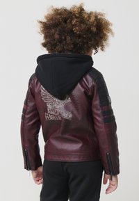 Bambino con capelli ricci che indossa una giacca di pelle bordeaux e nera con un'emblema a forma di ali e un cappuccio nero, in piedi di spalle.