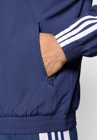 Modrý vetruodolný kabát má elastické manžety, zipsový vrecko na boku a tri biele pruhy na rukáve ako doplnkový detail.