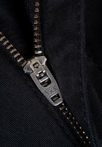 Schwarzer Stoff mit einem silbernen Metallreißverschluss, auf dem das sichtbare YKK-Logo zu sehen ist. Die Reißverschlusszähne sind gleichmäßig entlang des Schenkels verteilt, was auf hohe Qualität hinweist.