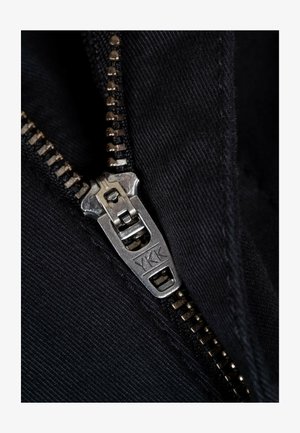 Schwarzer Stoff mit einem silbernen Metallreißverschluss, auf dem das sichtbare YKK-Logo zu sehen ist. Die Reißverschlusszähne sind gleichmäßig entlang des Schenkels verteilt, was auf hohe Qualität hinweist.
