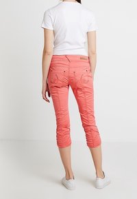Pantalones recortados color coral con una textura suave, detalles fruncidos en el dobladillo y bolsillos traseros bordados. Llevados con una camiseta blanca sencilla.
