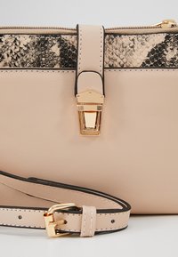 Beige kožená kabelka s texturovaným černým hadím vzorem, zlatými kovovými doplňky a nastavitelným popruhem, která má bezpečné zapínání.