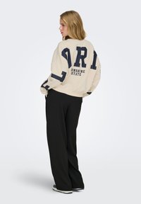 Jeune femme debout, portant un sweat-shirt beige avec un grand texte noir « FLORIDA SUNSHINE STATE » au dos, un pantalon large noir et des baskets.