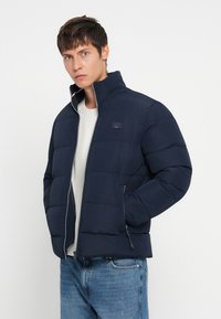 Young man wearing a navy blue padded jacket over a white knit sweater and blue jeans, standing with one hand in his pocket.