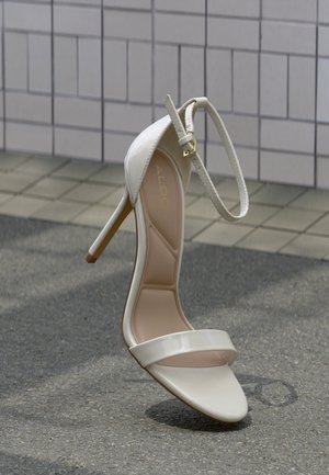 Riemchensandalette mit hohem Absatz aus glänzendem weißem Material. Verfügt über einen dünnen Knöchelriemen und zwei vordere Bänder, mit beigem Innenfutter und schlankem Stiletto-Absatz.