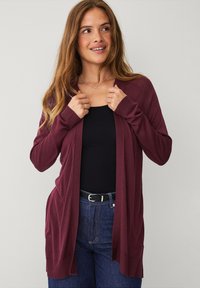 Bordeaux cardigan van zachte gebreide stof, met een open voorkant, ribbeltextuur langs de mouwen en onderkant, gedragen over een zwarte top en blauwe jeans.