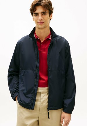 Ung mand iført en marineblå jakke med lynlås over en rød polo og beige bukser, stående mod en ensfarvet hvid baggrund.