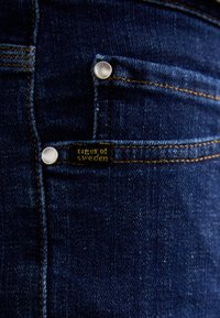Ciemnoniebieskie dżinsy z widocznym przeszyciem, małą przednią kieszonką oraz metką z logo "tiger of sweden" przy srebrnym guziku.