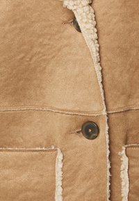 Giacca in suede beige con una fodera in pile crema testurizzato lungo il bordo. Presenta due tasche frontali e un bottone scuro per la chiusura.
