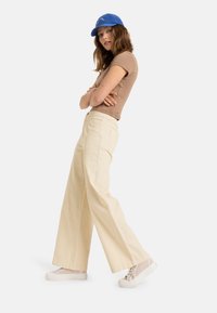 Beige ribbede vidde bukser med sidelommer, parret med en tætsiddende brun top og en blå kasket samt lyse sneakers.