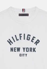 Weißes Baumwoll-T-Shirt mit Rundhalsausschnitt, das in dunkler blauer Schrift "HILFIGER NEW YORK CITY" aufgedruckt hat und ein rot-blau gestreiftes Etikett im Nacken trägt.