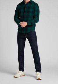 Groen geruit flanellen overhemd met knoop-omlaag kraag, gecombineerd met donkerblauwe spijkerbroek en witte sneakers met tan accenten.