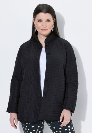 Femme aux longs cheveux foncés portant une veste matelassée noire sur un haut blanc et un pantalon noir à pois, debout devant un fond blanc.