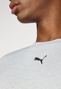 Grå bomull T-skjorte med en svart Puma-logo på øvre venstre side, rund hals og myk tekstur.