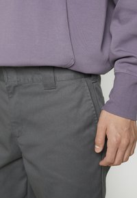 Main reposant à côté d'un pantalon gris, portant une chemise lavande à manches longues rentrée à la taille.