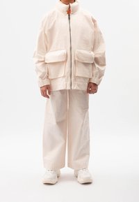 Béžová bomber bunda s vysokým límcem, dvěma předními kapsami a elastickými manžetami. Kombinována s širokými kalhotami s plisováním ve stejné barvě.