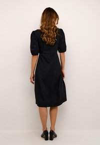 Robe noire midi avec des manches bouffantes, une taille cintrée et des détails de couture subtils. Texture lisse, longueur mi-mollet, assortie à des talons noirs.