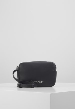 Black Calvin Klein leather wristlet with silver logo and adjustable strap placed on white surface against gray background.