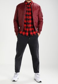 Man som bär en röd bomberjacka, röd och svart ruta skjorta, svarta byxor och vita sneakers, står med händerna i fickorna.