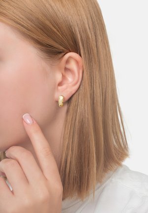 Nærbillede af en kvinde med glat blond hår, iført små teksturerede guldøreringe og berører sin kind med en velplejet finger.