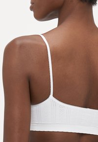 Hvit bralette med tynne stropper, teksturert stoff med et subtilt mønster, og delikat bølget kanting langs kantene.