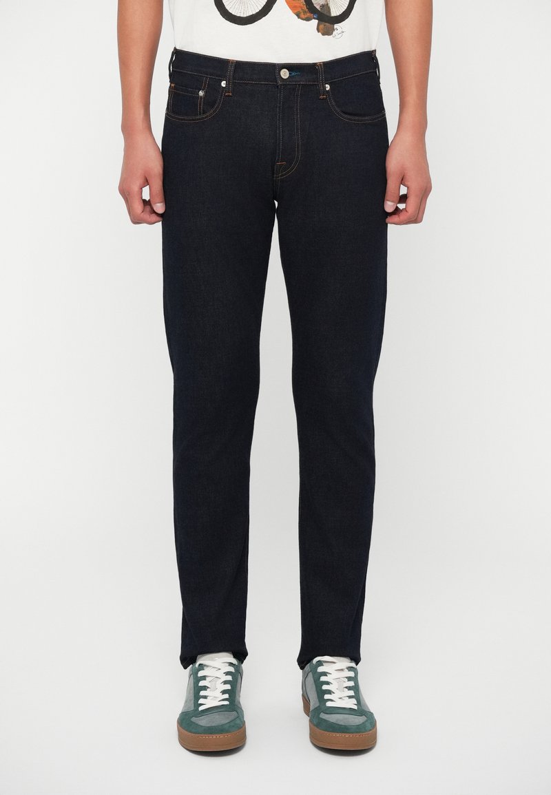 Jeans de ganga azul escuro com um corte slim, apresentando costuras em contraste, design de cinco bolsos e fecho de botão. Combinados com ténis verdes e cinzentos.
