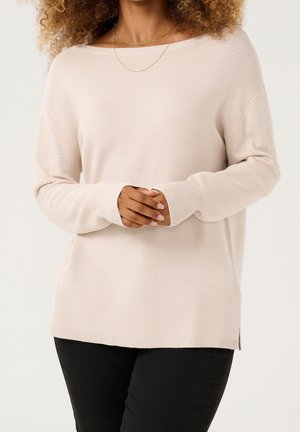 Light beige ribbed knit sweater with a wide neckline, long sleeves, and side slits at the hem, paired with black pants.