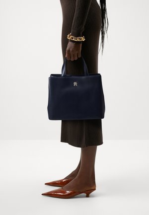 Navy leather tote bag with structured shape, two top handles, and a gold logo. Worn with a brown ribbed dress and pointed brown heels.