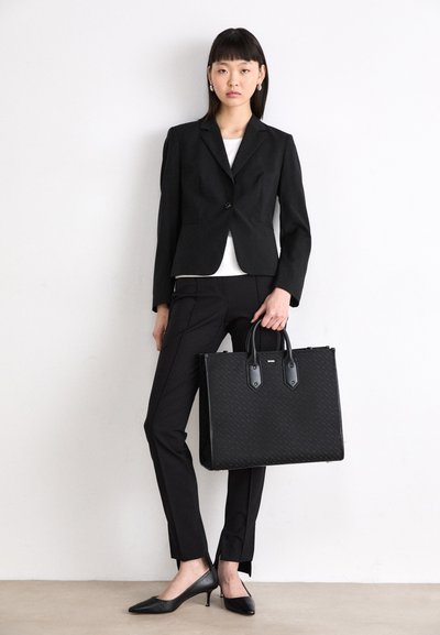 Sac à main noir structuré avec motif texturé, doté de deux poignées et d'une quincaillerie noire contrastée. Modèle portant un blazer et un pantalon noirs.