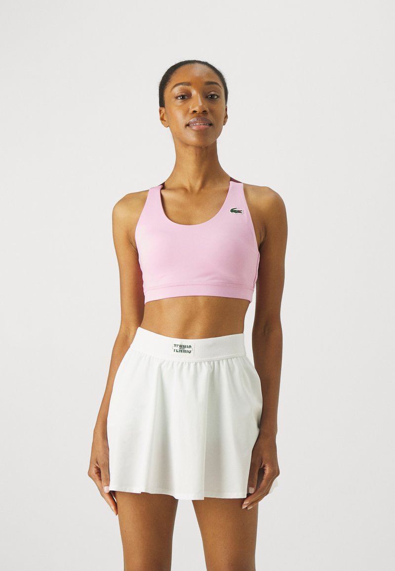 Sostén deportivo rosa con escote redondo y tirantes negros, combinado con una falda blanca de pliegues que cuenta con una cintura elástica y un detalle de logo.