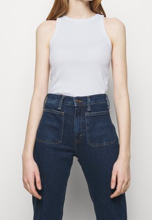 Vrouw draagt een mouwloze witte top en donkerblauwe jeans met hoge taille en voorzakken, staand tegen een effen lichte achtergrond.