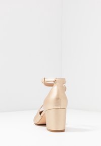 Goldfarbener Schuh mit niedrigem Blockabsatz und Knöchelriemen sowie Schnalle, von hinten auf einer weißen Oberfläche mit schlichtem weißen Hintergrund gezeigt.