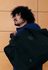 Svart duffelbag med glatt tekstur, stor hovedrom, sidelomme med glidelås og preget logo. Bæres med svarte stropper.