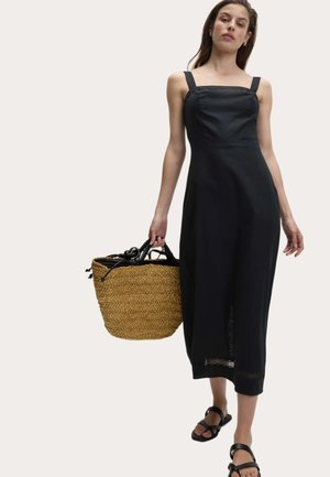 Kvinde iført en ærmeløs sort midi-kjole og sorte sandaler, bærende på en stor vævet stråtaske med sorte håndtag.