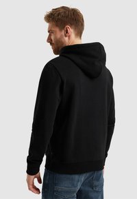 Felpa nera in cotone con cappuccio, polsini e orlo a costine. La vista da dietro mostra una vestibilità rilassata e un design minimalista. Jeans in denim visibili sotto.