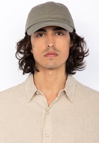 Khaki baseballpet met een gebogen rand, eenvoudig ontwerp en geborduurd merk. Draagt bij een lichtbeige overhemdbloes.