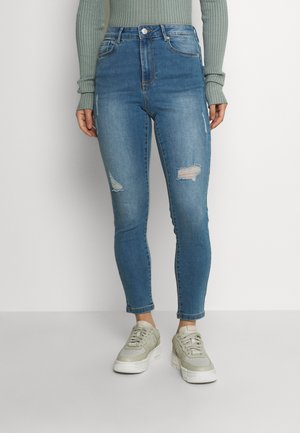 Kvinde iført lyseblå, slidte skinny jeans, salviegrøn ribbet langærmet top og beige sneakers, stående mod en hvid baggrund.